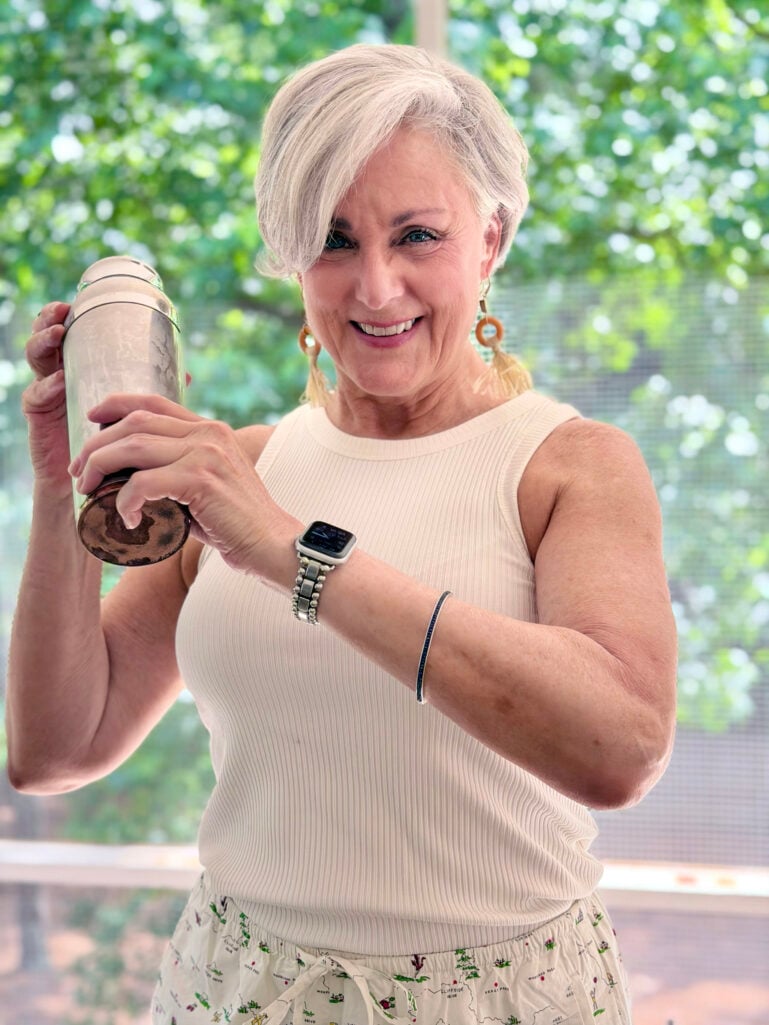 Beth in a cream ribbed tank top and tassel earrings smiling while shaking a silver cocktail shaker on the porch, with green foliage behind her.