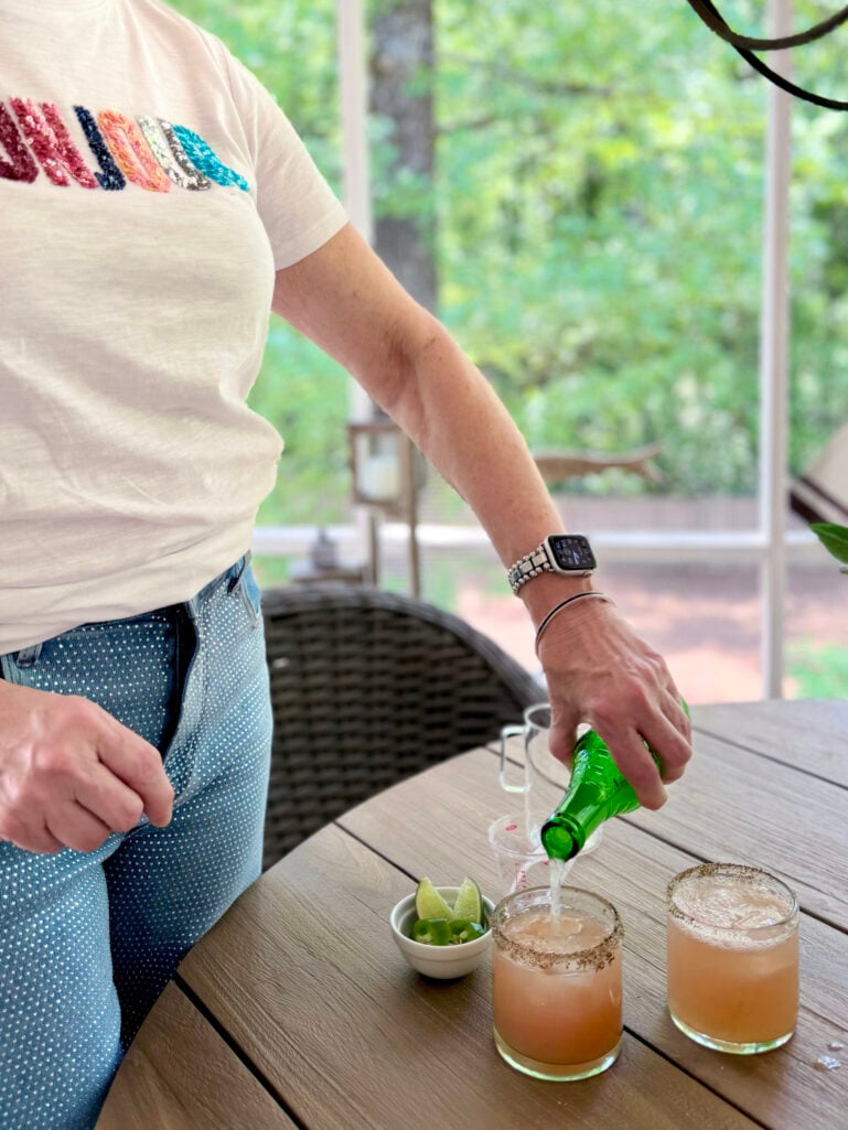 Beth leans over an outdoor table pouring Jarritos Grapefruit soda into two tajín-rimmed rocks glasses filled with spicy paloma, a small white bowl of jalapeños and lime wedges nearby, the screened porch and garden visible behind her.