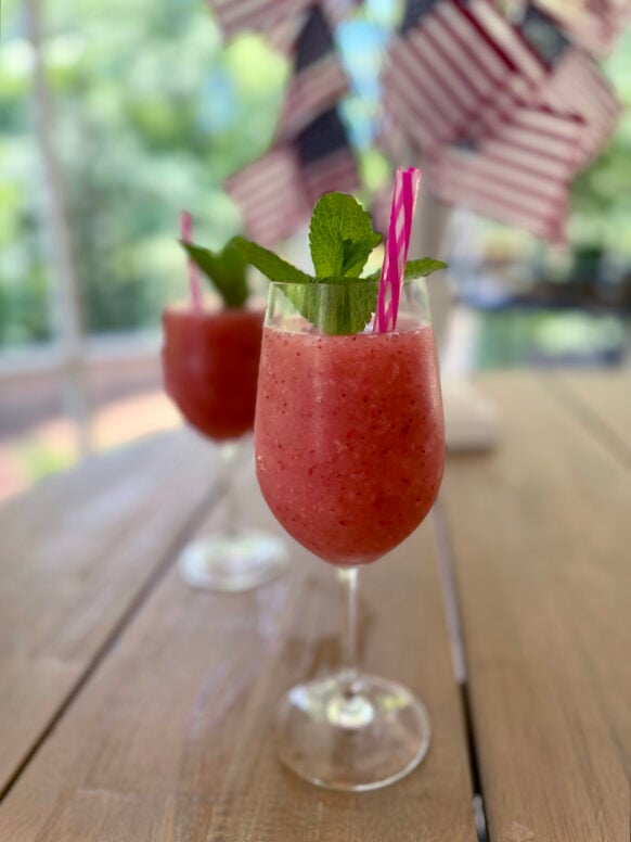 Close-up of two frozen strawberry frosé cocktails in stemmed glasses with fresh mint and pink-and-white striped straws, on a wood table with American flags softly blurred behind.