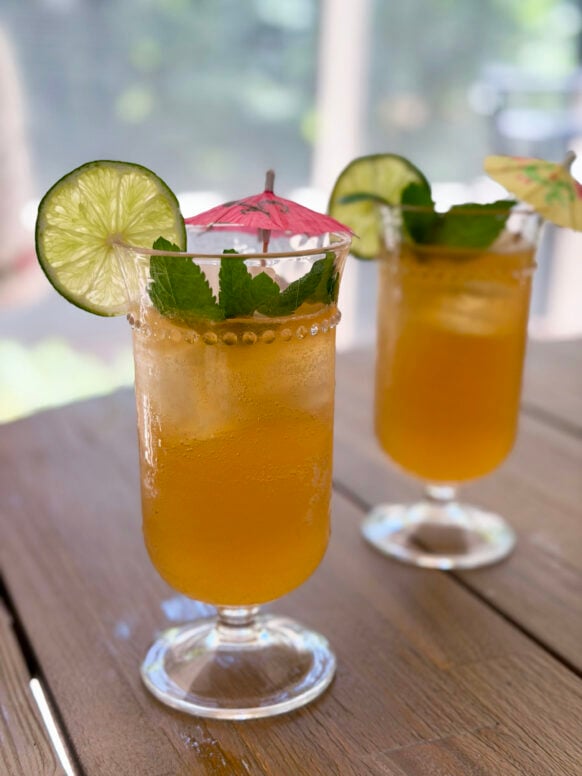 Close-up of two tropical rum cocktails in stemmed hurricane glasses with lime wheel garnishes, fresh mint sprigs, and pink paper umbrellas, on a wood patio table.