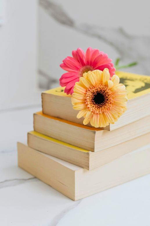 Stack of neutral-toned books topped with bright pink and yellow gerbera daisies on a white marble surface, creating a cheerful and cozy reading-inspired flat lay.