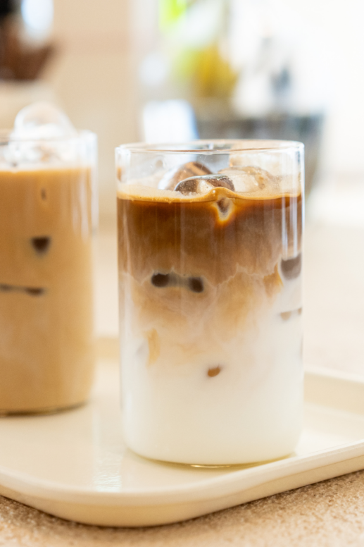 Close-up of layered iced coffee in a clear glass tumbler with milk swirling into espresso over ice cubes, styled on a neutral tray with a second iced latte softly blurred in the background.