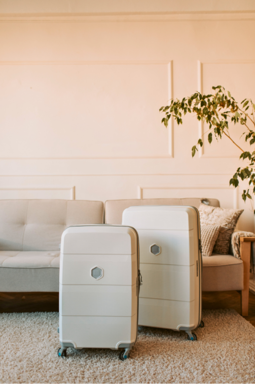 Two cream-colored hard-shell spinner suitcases styled in a warm neutral living room with a beige sofa, textured rug, and indoor tree, creating a calm and elevated travel scene.