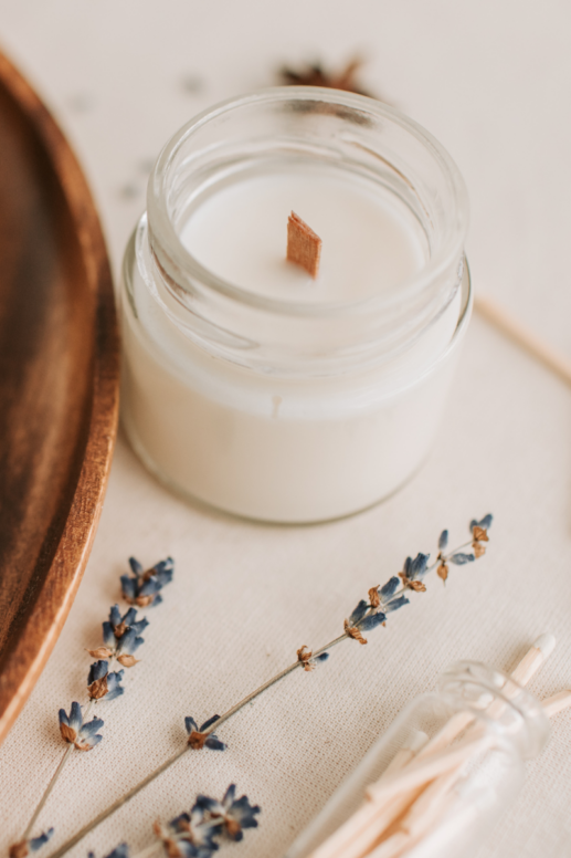 Soft white candle in a clear glass jar with a wooden wick, styled on neutral linen fabric beside dried lavender sprigs, a wooden tray, and glass match holder for a cozy spa-inspired setting.