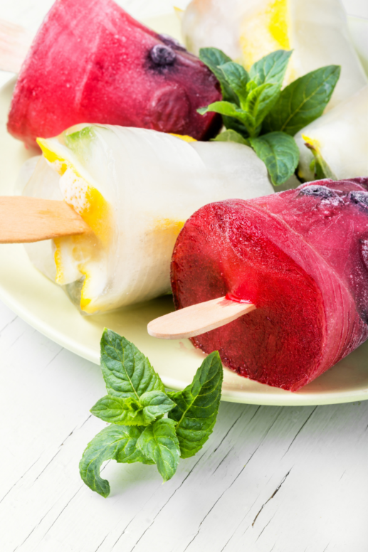 Colorful homemade fruit popsicles made with creamy citrus and mixed berry layers, styled on a white plate with fresh mint leaves and ice cubes on a rustic white wood surface.