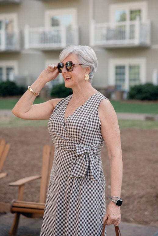 Woman with short silver hair wearing oversized sunglasses, statement gold earrings, and layered gold bracelets with a sleeveless gingham wrap dress.