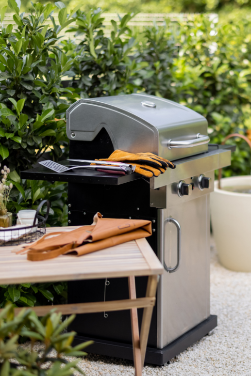Modern outdoor grilling station with stainless steel gas grill, leather apron, grilling tools, and barbecue gloves set against lush backyard greenery and patio décor.