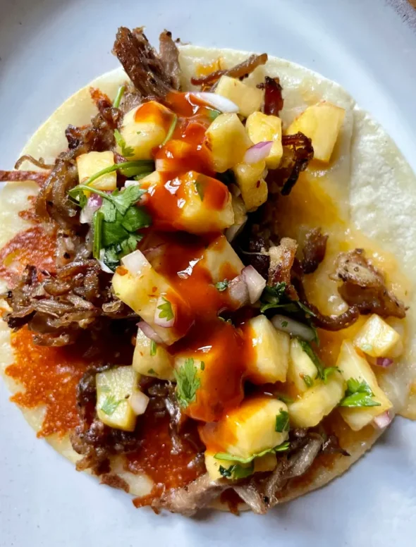 Close-up overhead of a single finished pork taco on a white plate topped with chipotle pineapple salsa and a drizzle of hot sauce over the crispy cheese-crusted corn tortilla