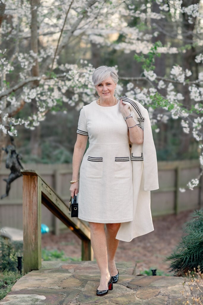 Woman over 50 with silver hair wearing a cream cotton bouclé short-sleeve shift dress with a matching cream duster coat draped over her arm, styled with black patent Louboutin pumps, a gold chain link necklace, gold cuff bracelet, and a small black structured top-handle bag — an elegant monochromatic cream outfit idea for spring or summer occasions for women over 50.