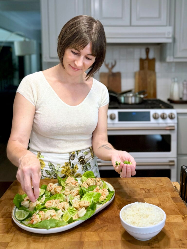 Kelly in a cream cashmere tee and floral wide-leg pants garnishing a platter of butter lettuce cups filled with shrimp on a butcher-block kitchen island, with a small bowl of jasmine rice nearby.