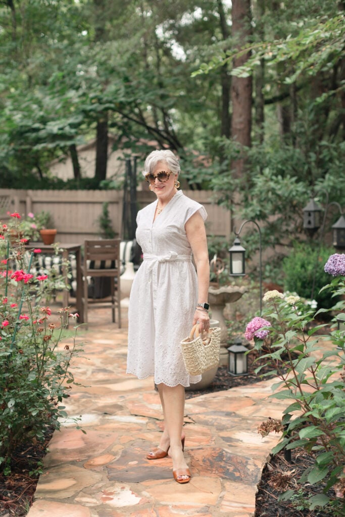Woman over 50 with silver hair wearing a white eyelet midi shirtdress with a tie waist, tortoise sunglasses, gold drop earrings, and cognac leather slide sandals, carrying a woven straw basket bag in a flagstone garden with roses and hydrangeas — a summer white dress outfit idea for women over 50.