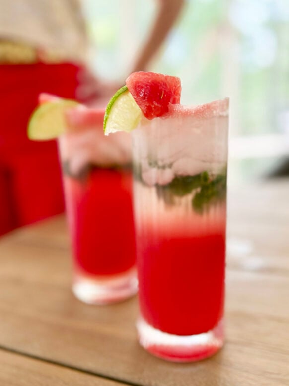 Two tall highball glasses filled with vibrant red watermelon mojitos sit on an outdoor wooden table, each garnished with a fresh watermelon chunk and lime wheel on the rim, muddled mint visible through the glass, Beth's red jeans softly blurred in the background.