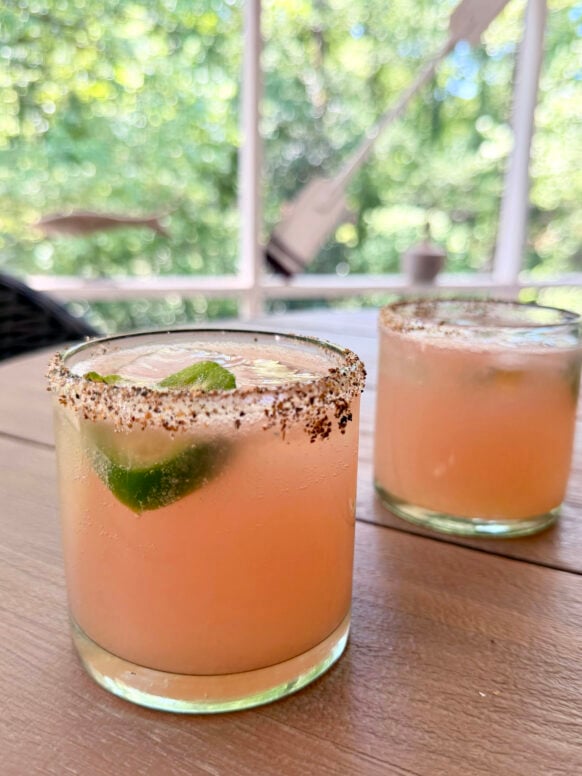 Two spicy paloma cocktails in tajín-rimmed rocks glasses sit on a wooden outdoor table, each garnished with a fresh jalapeño slice, with lush green bokeh trees visible through the screened porch behind them.