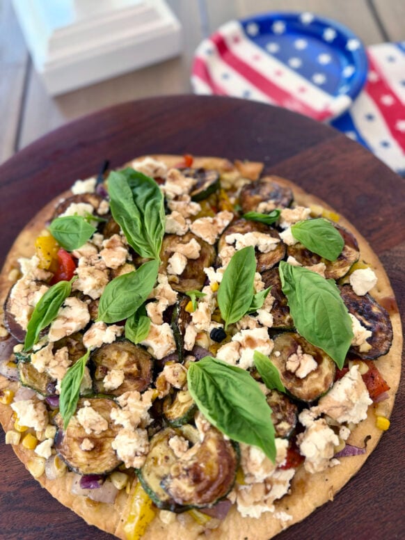 Finished grilled veggie flatbread topped with charred zucchini, peppers, corn, red onion, crumbled goat cheese, fresh basil, and a balsamic drizzle, on a round wood board with a stars-and-stripes dish in the background.
