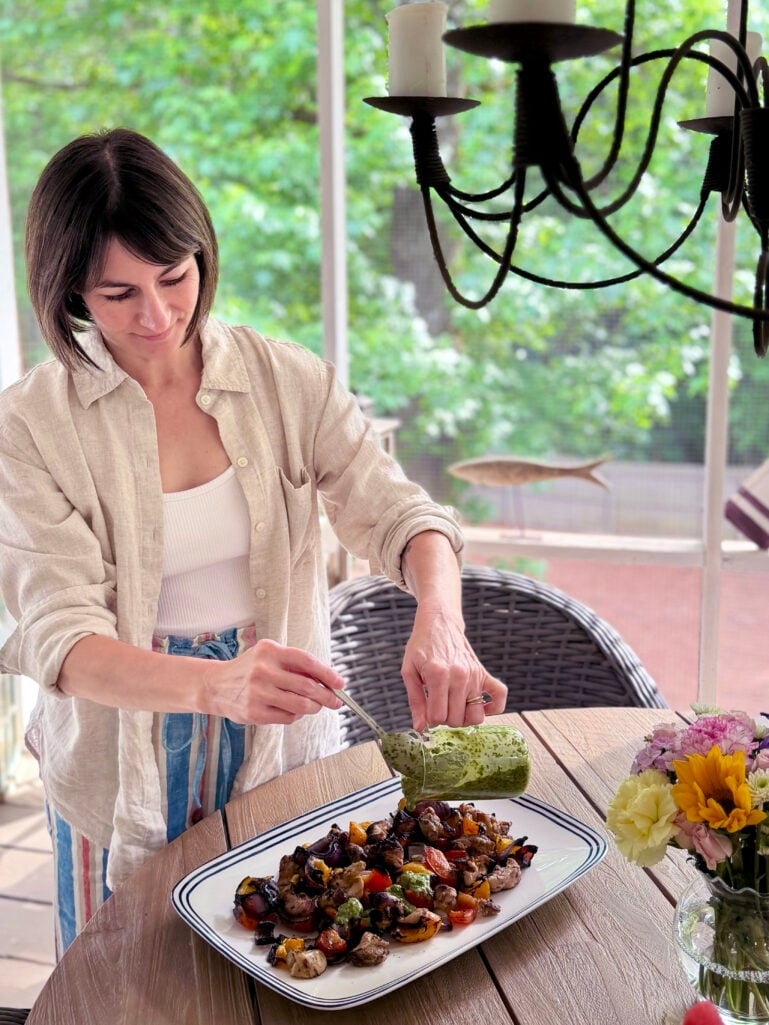 Kelly stands at the outdoor porch dining table, drizzling bright green chimichurri sauce from a small squeeze bottle over a platter of grilled chicken and vegetables, wearing her natural linen overshirt open over a white tank and colorful striped linen pants, a mason jar of summer flowers beside the platter.