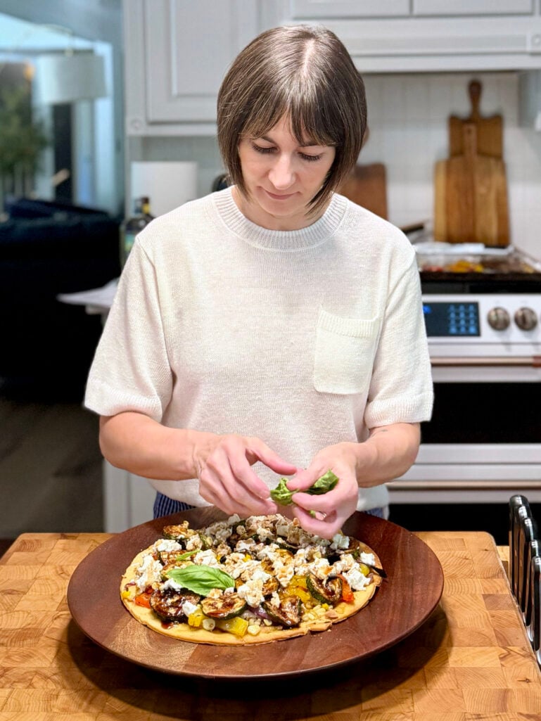 Kelly in a cream knit top tearing fresh basil over a grilled vegetable flatbread topped with zucchini, goat cheese, and balsamic, on a dark wood board atop a butcher-block kitchen island.