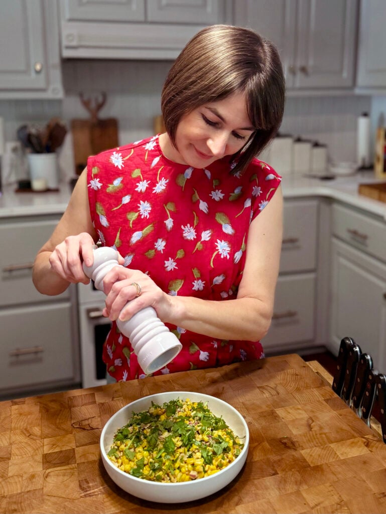 Kelly stands at a butcher block kitchen counter, smiling as she grinds fresh black pepper over a white bowl of street corn salad topped with fresh cilantro, wearing her vibrant red floral sleeveless top, with gray cabinets and kitchen tools visible behind her.