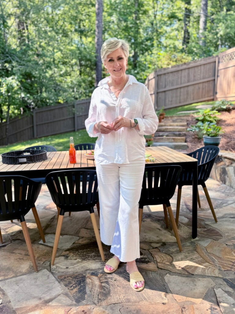 Woman over 50 with silver hair wearing a white linen button-down shirt with white wide-leg cropped denim jeans and raffia woven slide sandals on a flagstone patio with a teak dining table — a mixed-texture all-white outfit idea for women over 50.