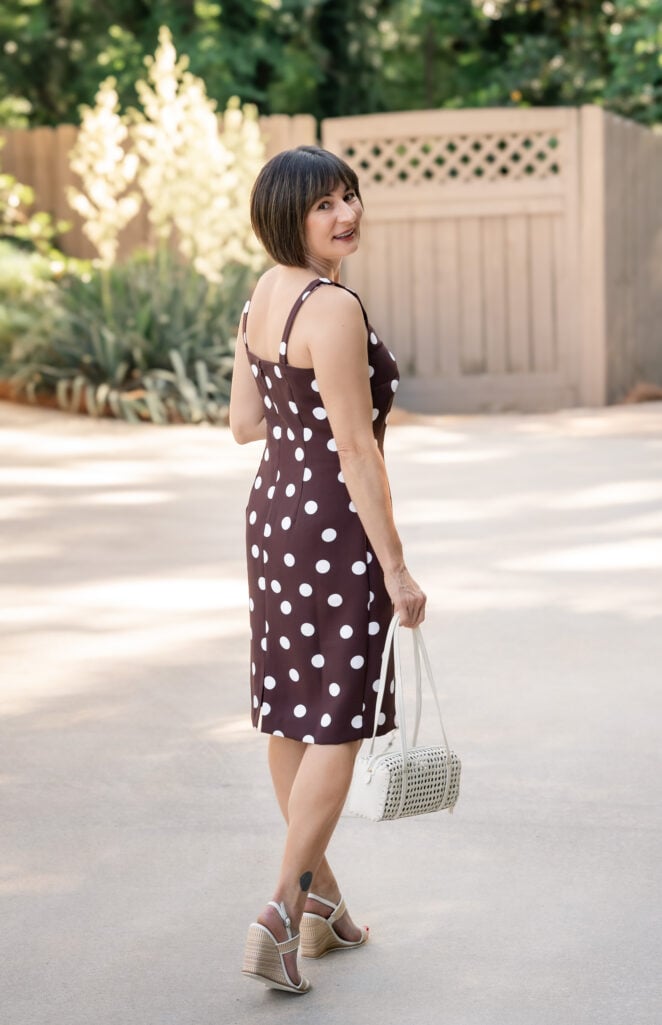 Back view of Kelly in brown polka dot sheath dress showing the square neckline straps and knee-length hem, holding cream woven cane shoulder bag — petite summer dress fit detail