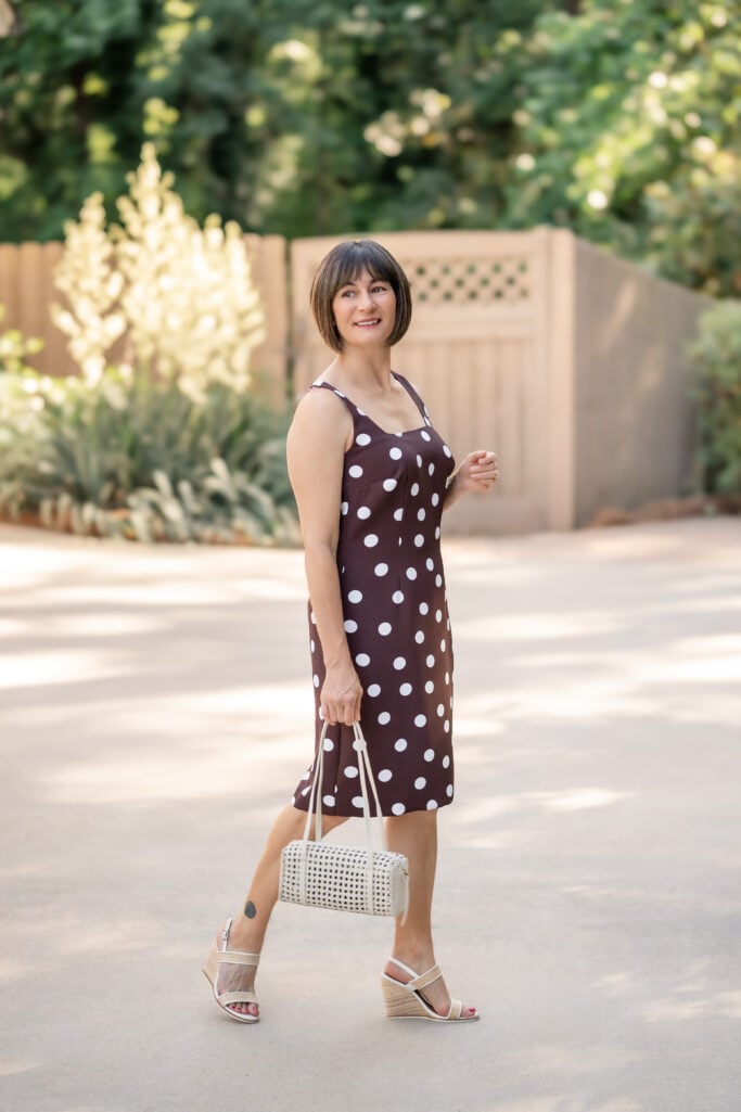 Kelly standing in profile wearing brown and white polka dot sheath dress with raffia wedge sandals and cream cane bag — petite summer sheath dress styling