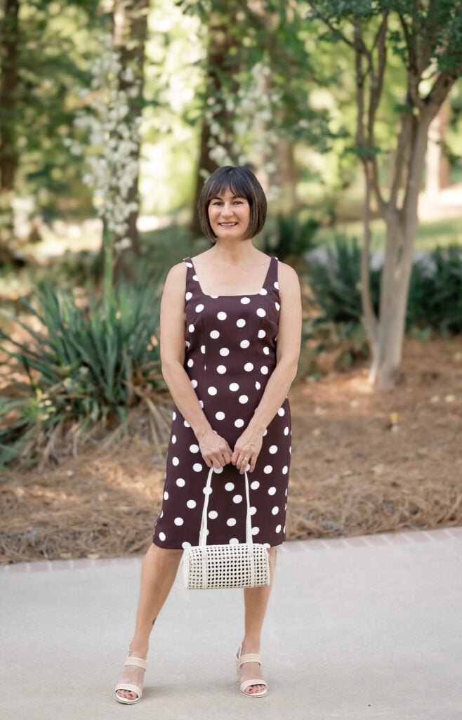 Kelly facing forward in chocolate brown polka dot square neck sheath dress holding cream cane shoulder bag — best petite summer dress that fits without alterations