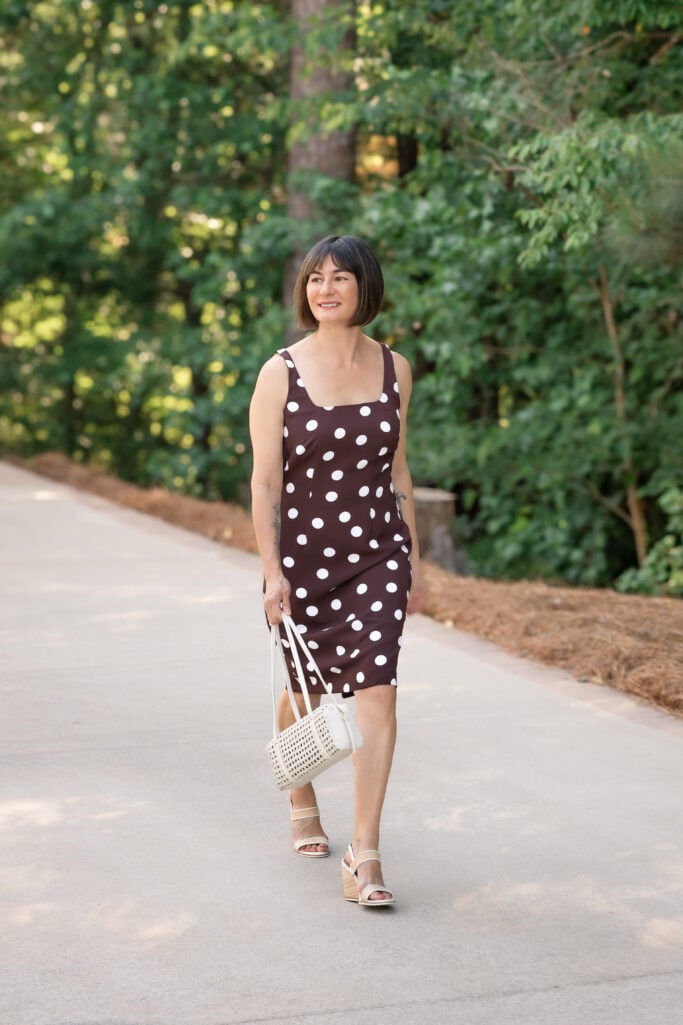 Kelly walking toward camera in a brown and white polka dot sheath dress with cream cane bag and raffia wedges — petite summer sheath dress styled for daytime
