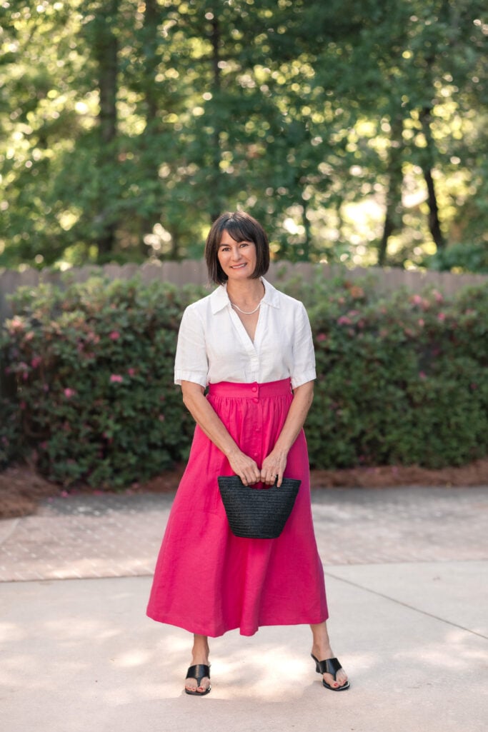 Kelly in full-length petite summer outfit featuring white linen popover top tucked into hot pink button-front linen maxi skirt with black wedge sandals and straw bag