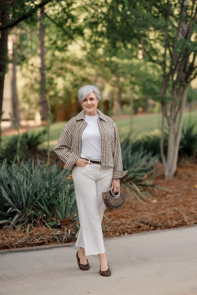 Woman over 50 with silver hair wearing a brown and white gingham canvas barn jacket over a white tee with cream wide-leg cropped jeans, a thin brown belt, brown woven leather flats, and a tortoise-handled woven straw handbag in a wooded garden — a polished casual summer outfit idea featuring white wide-leg pants for women over 50.