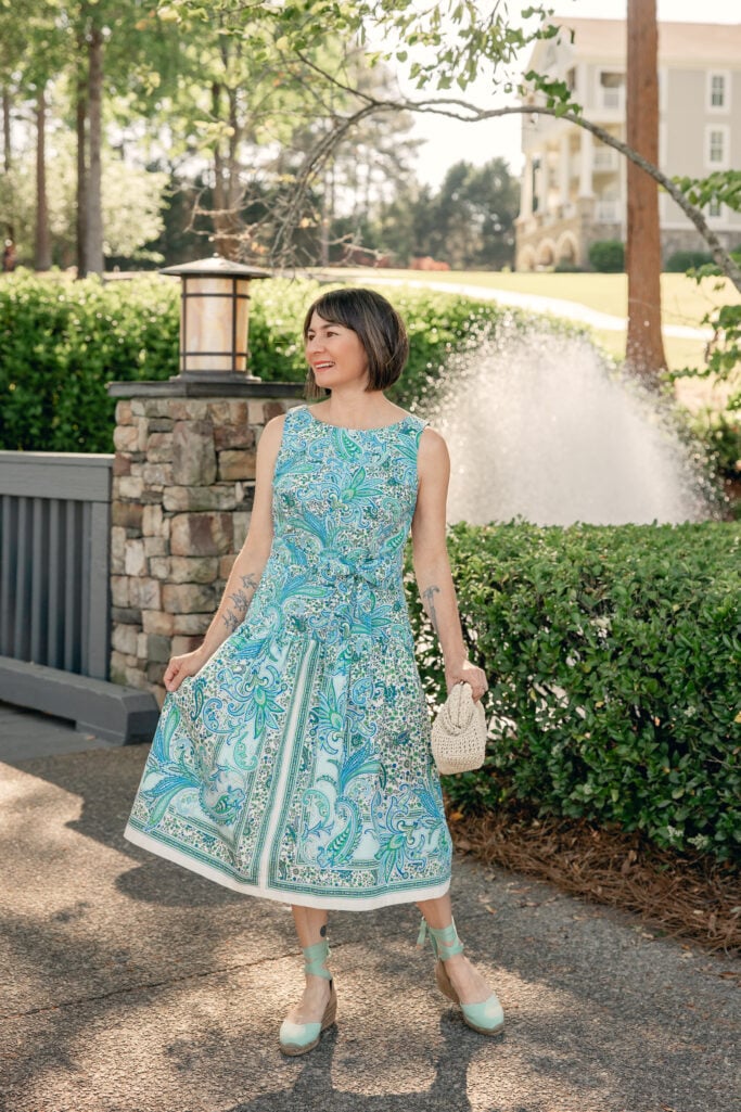 Woman with a dark bob wearing a blue and green paisley fit and flare midi dress with a self-tie waist and white hem border, carrying a small cream woven clutch and wearing mint green lace-up wedge espadrilles — a full-length view of the Mother's Day dress in petite sizing showing the hem length and silhouette.