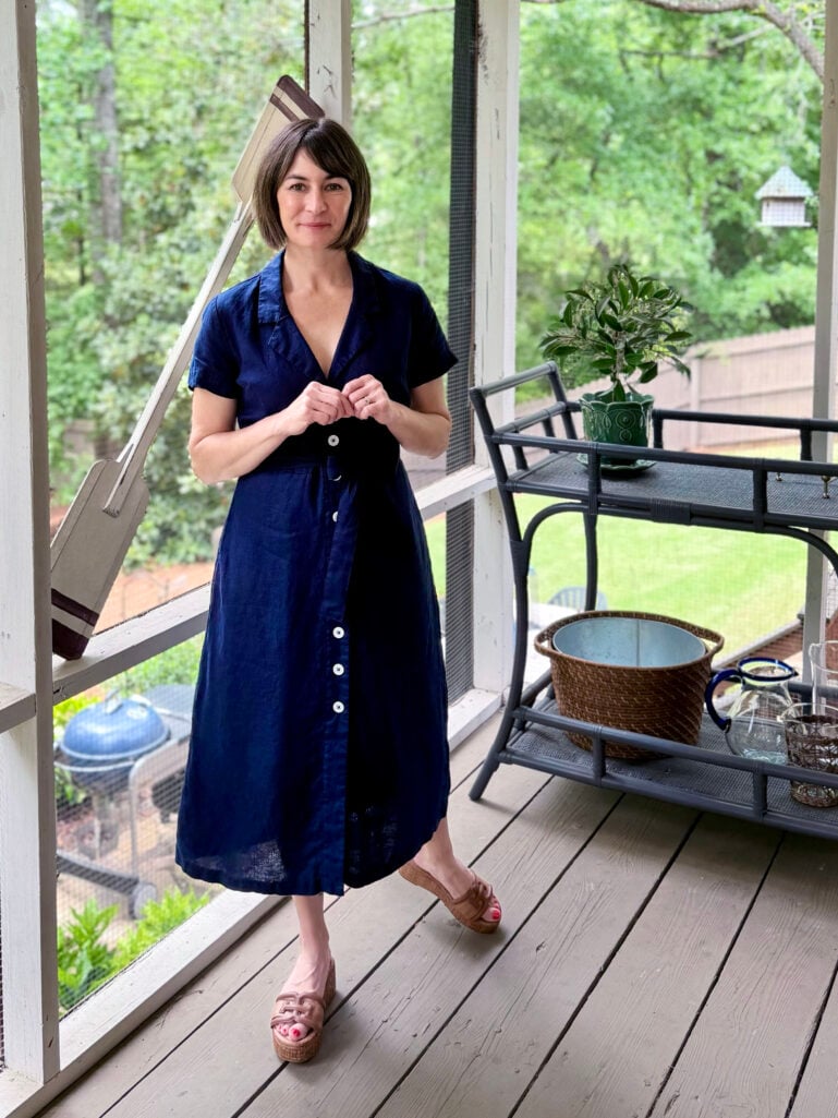 Kelly stands on the screened porch in a navy European linen button-front shirtdress with a wide woven leather belt and espadrille wedge sandals, hands lightly clasped in front, with a bar cart and green backyard visible behind her.