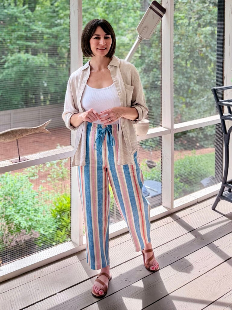 Kelly stands on the screened porch in a natural linen Garçon button-up shirt worn open over a white rib scoop neck tank, with wide-leg colorful striped linen crop pants tied at the waist and brown toe loop sandals, hands lightly clasped in front, smiling softly at the camera.