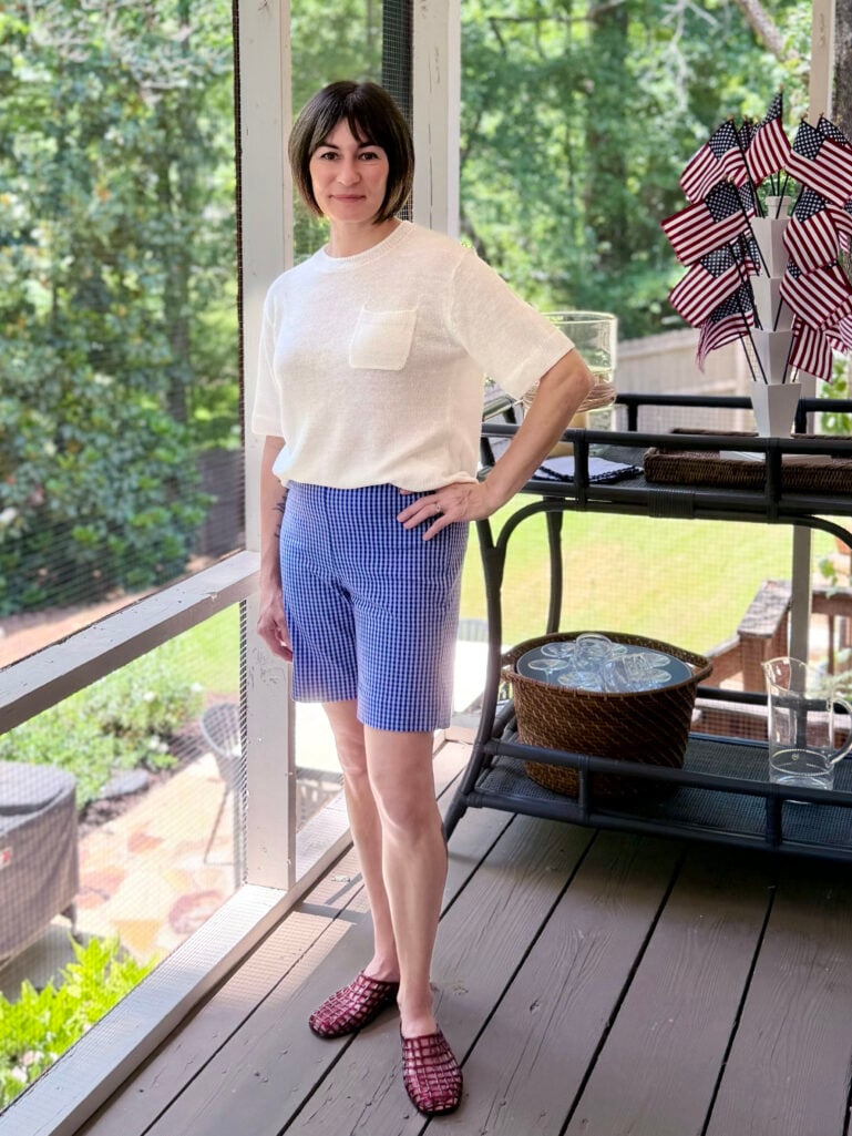 Full-length view of Kelly on the screened porch in a cream pocket sweater tee, blue gingham Bermuda shorts, and berry-red netted jelly mules, hand on hip, with American flags and a bar cart nearby.