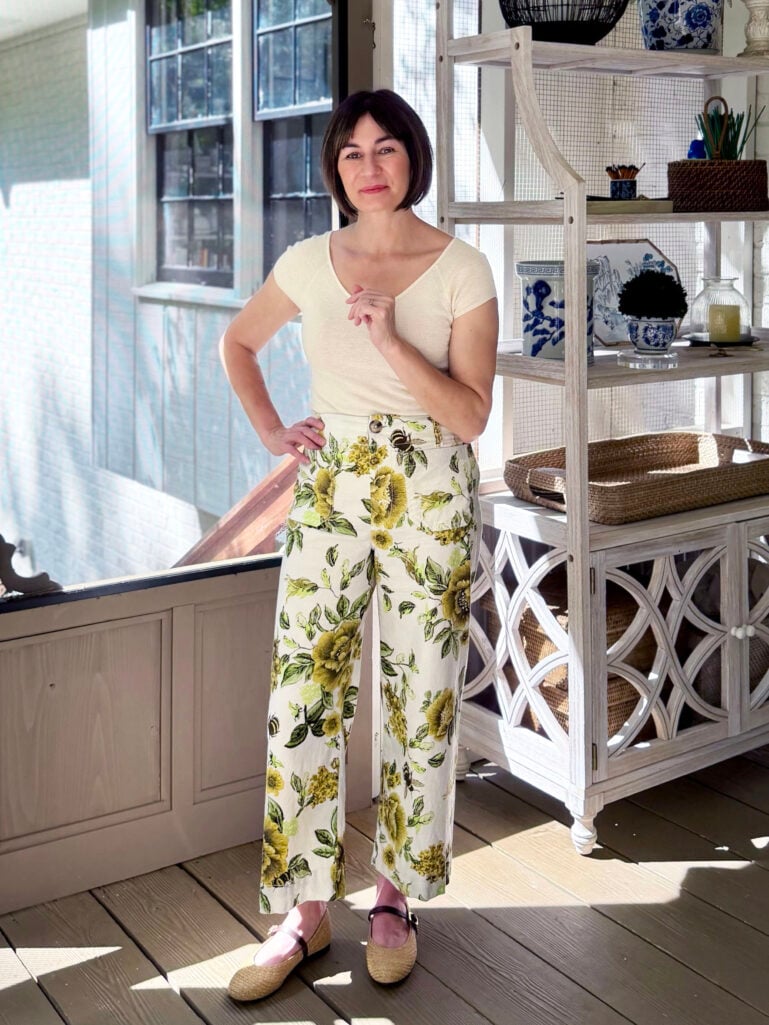 Kelly standing on the screened porch in a cream cashmere tee and green-and-yellow floral wide-leg linen-blend pants with raffia Mary Jane flats, hand on hip, with a window and baker's rack behind her.