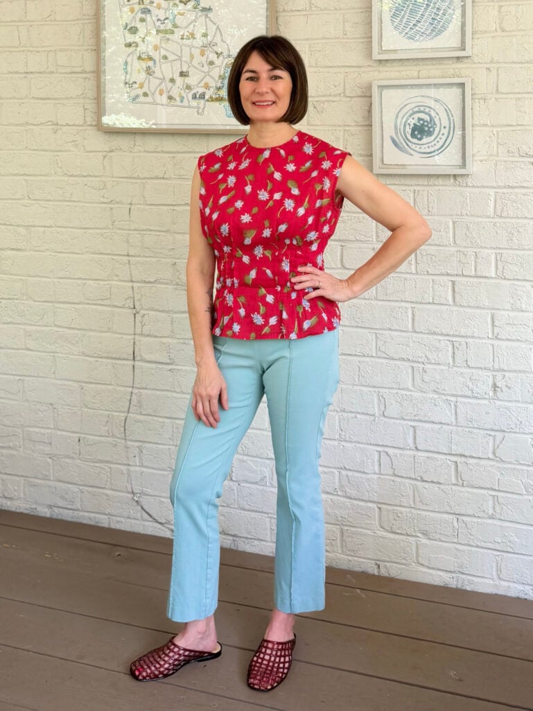 Kelly stands on the porch against a white brick wall, one hand on her hip, wearing a vibrant red floral sleeveless peplum top, aqua straight-leg Ivy pants with a front seam detail, and burgundy netted jelly mule sandals.