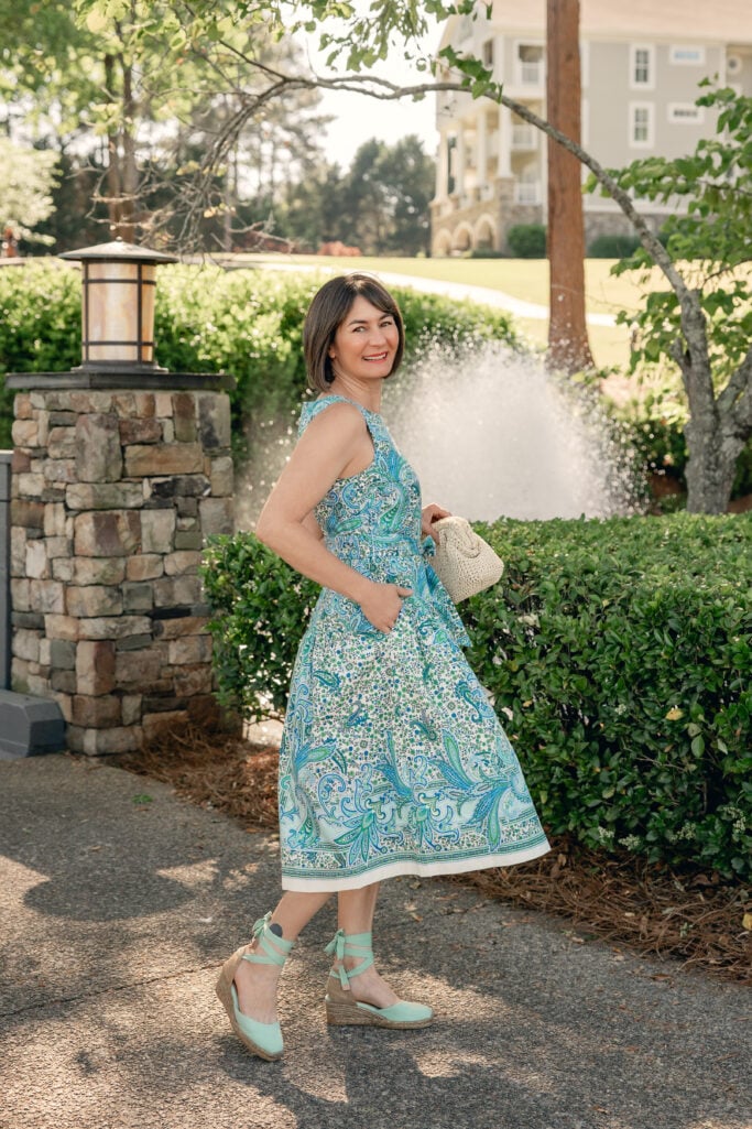 Woman with a dark bob smiling over her shoulder outdoors wearing a blue and green paisley fit and flare midi dress, holding a small cream woven clutch and wearing mint ankle-strap wedge espadrilles — a back view showing the movement of the Mother's Day dress in petite sizing.