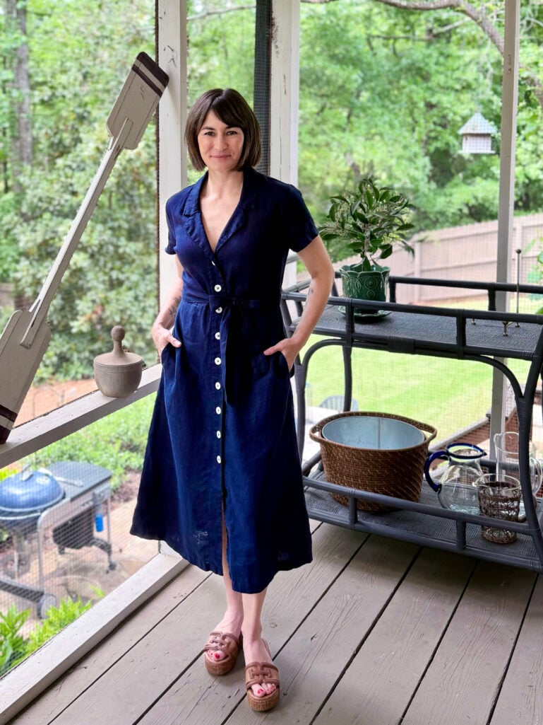 Full-length view of Kelly on the screened porch in her navy linen shirtdress belted at the waist, both hands in her pockets, one foot slightly crossed, smiling at the camera with lush green trees and porch details behind her.