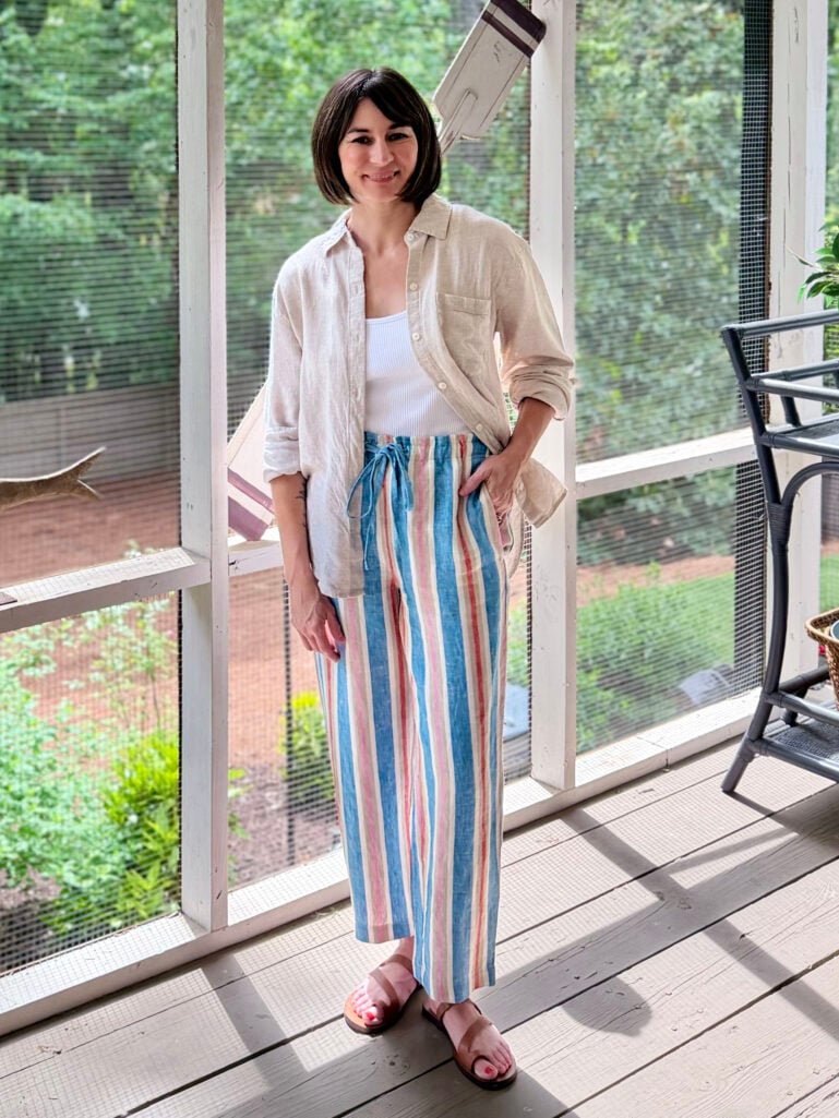 Full-length view of Kelly on the screened porch in her linen overshirt, white tank, and multicolor wide-leg striped linen pants with brown toe loop sandals, one hand in her pocket, smiling at the camera with the porch and green trees behind her.