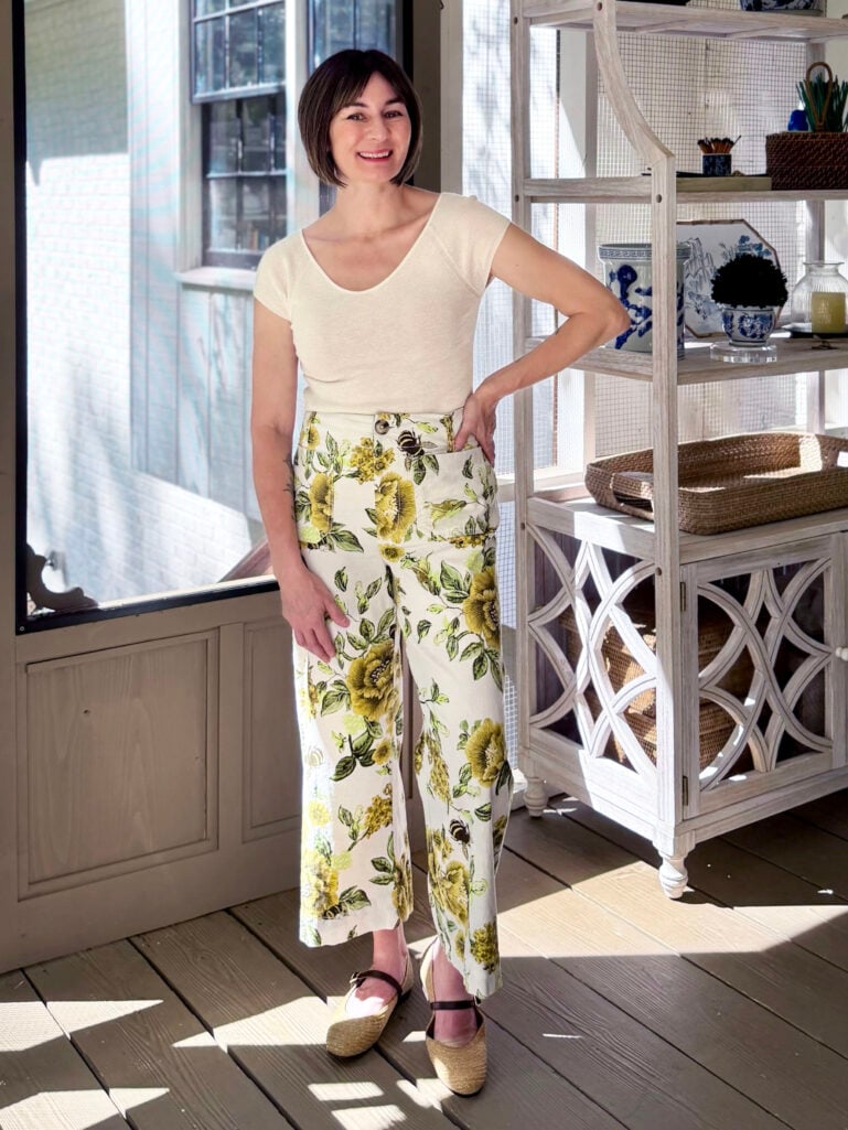 Full-length view of Kelly on the screened porch in a cream cashmere tee and floral wide-leg pants with raffia flats, smiling with windows and a baker's rack behind her.