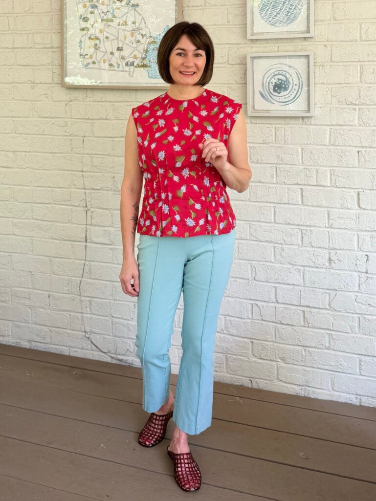 Full-length view of Kelly on the porch in her red floral peplum top, aqua Ivy pants, and burgundy jelly mules, smiling with one hand raised lightly at her side, the Georgia map print and blue abstract art visible on the white brick wall behind her.