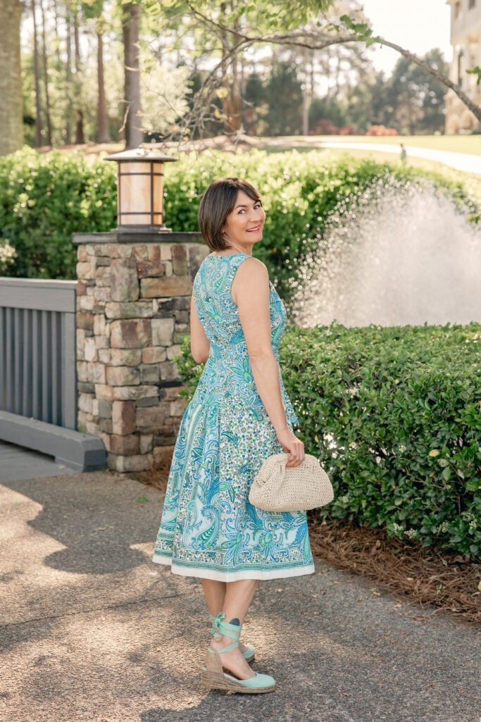 Woman with a dark bob laughing and looking to the side outdoors in a blue and green paisley fit and flare midi dress with a self-tie waist, holding a small cream woven clutch and wearing mint ankle-strap wedge espadrilles — a joyful candid shot of the Mother's Day dress in petite sizing.