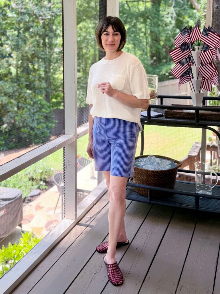 Petite woman over 50 with brunette bob wearing blue and white gingham Bermuda shorts with a relaxed cream pocket sweater tee and oxblood netted jelly mules — a classic gingham Bermuda shorts outfit idea for petite women over 50.