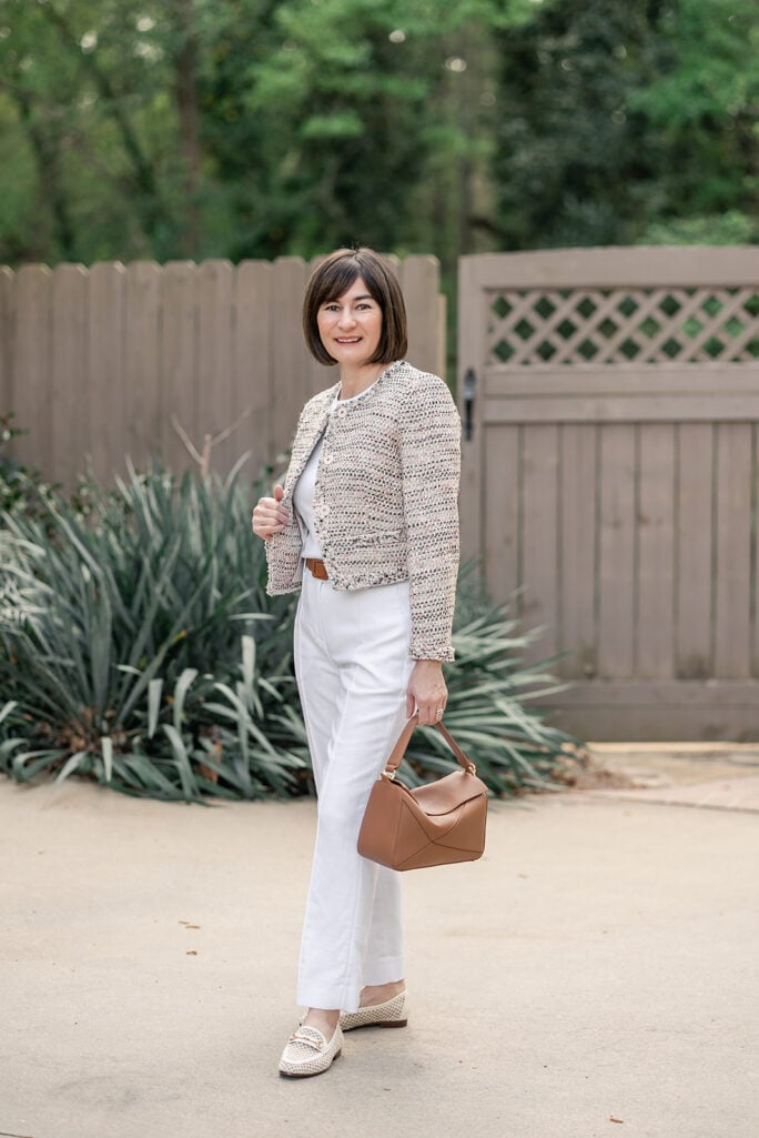 Woman wearing a cream and multicolor tweed cropped jacket over a white tee with white linen-blend wide-leg pants, woven beige loafers, and a structured cognac top-handle handbag in a garden setting — a transitional spring-to-summer outfit idea featuring white linen pants for women over 50.