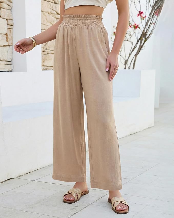 Tan linen-blend wide-leg pull-on pants with a smocked paper-bag elastic waistband, styled with a cream cropped top, gold bracelet, and natural woven slide sandals against a white stone wall — versatile pull-on linen pants for summer for women over 50.