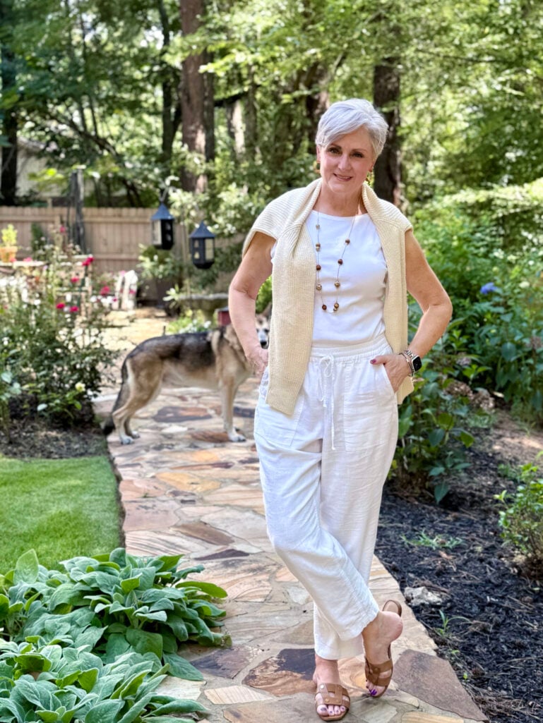 Woman over 50 with silver hair wearing white drawstring linen pants with a white short-sleeve tee, a cream knit cotton sweater draped over the shoulders, long wooden beaded necklace, and tan slide sandals on a flagstone garden path — an easy summer outfit idea featuring white linen pants for women over 50.