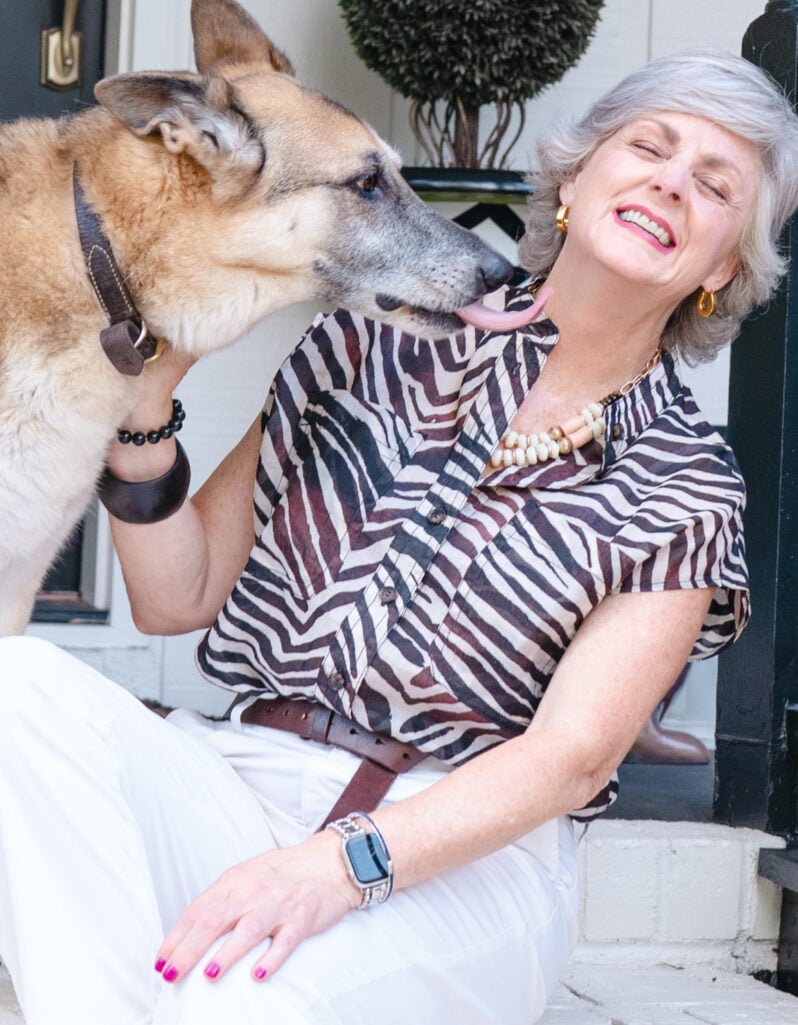 Beth sits on the front porch steps in 2022, laughing with her eyes closed and her head tilted back as Oscar, her large German Shepherd mix, enthusiastically licks her cheek with his tongue, Oscar wearing a brown leather collar, a lush green topiary visible on the porch behind them.