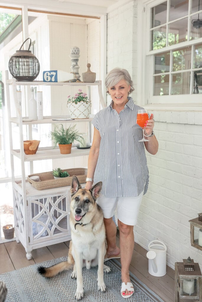 Beth stands on the screened porch in 2022, smiling and holding an orange cocktail, with Oscar — her large happy German Shepherd mix — sitting contentedly right beside her, the white porch shelving with decorative accessories and a window visible in the background behind them.