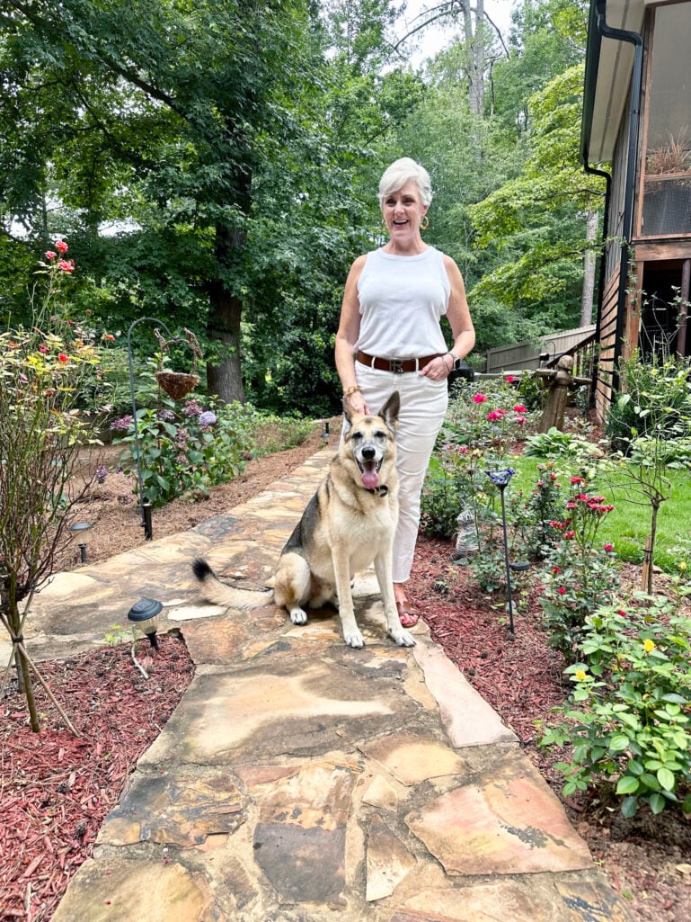 Beth stands on a stone garden path in 2023, laughing and resting her hand on Oscar, her large German Shepherd mix, who sits tall beside her with his tongue out and a big happy expression, red roses and lush garden plantings blooming on either side of the path behind them.