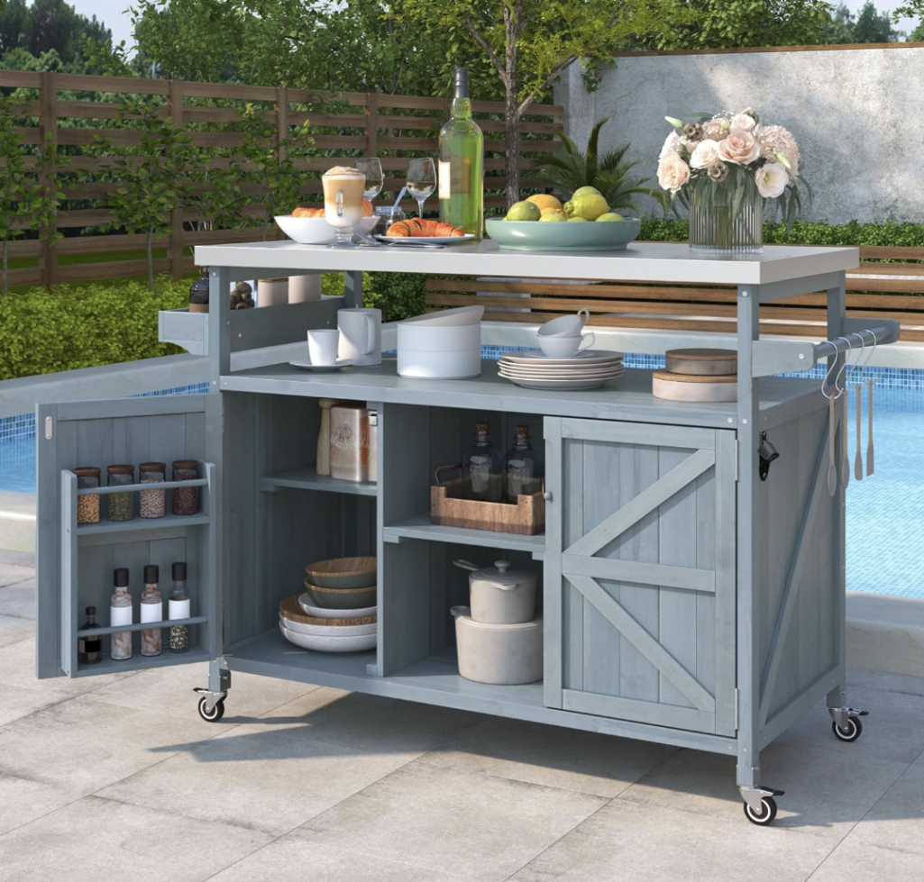Blue-gray outdoor grill cart with stainless steel top, open shelving, cabinet storage, spice rack, towel hooks, and rolling wheels styled beside a backyard pool patio.