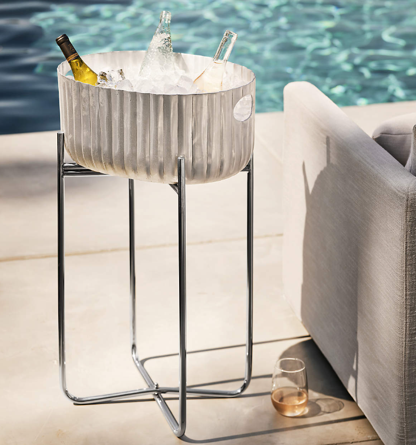 Silver fluted beverage tub on a stainless steel stand beside a swimming pool, filled with ice and chilled wine bottles next to outdoor lounge seating.