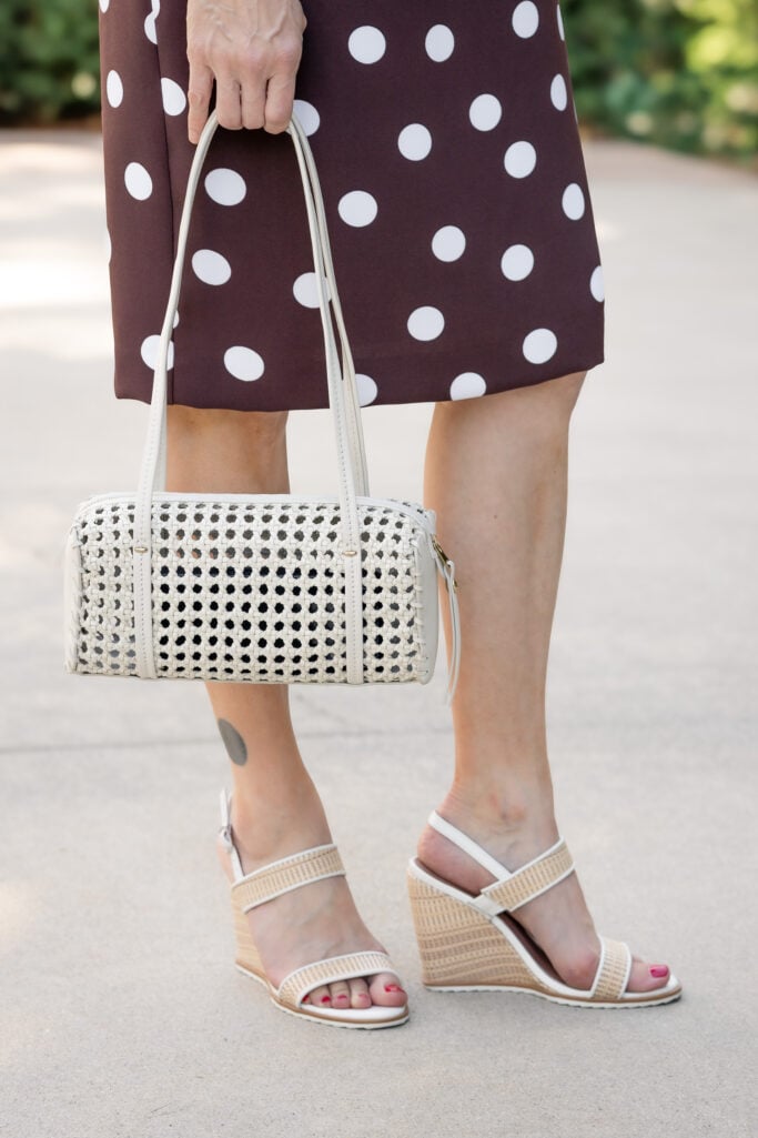 Close-up of cream woven cane shoulder bag and raffia and white wedge sandals styled with brown polka dot sheath dress — petite summer accessories detail